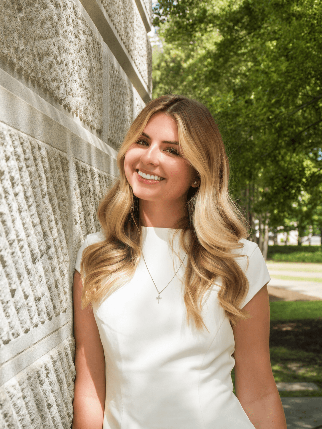 Graduate in a white dress smiling beside a stone wall